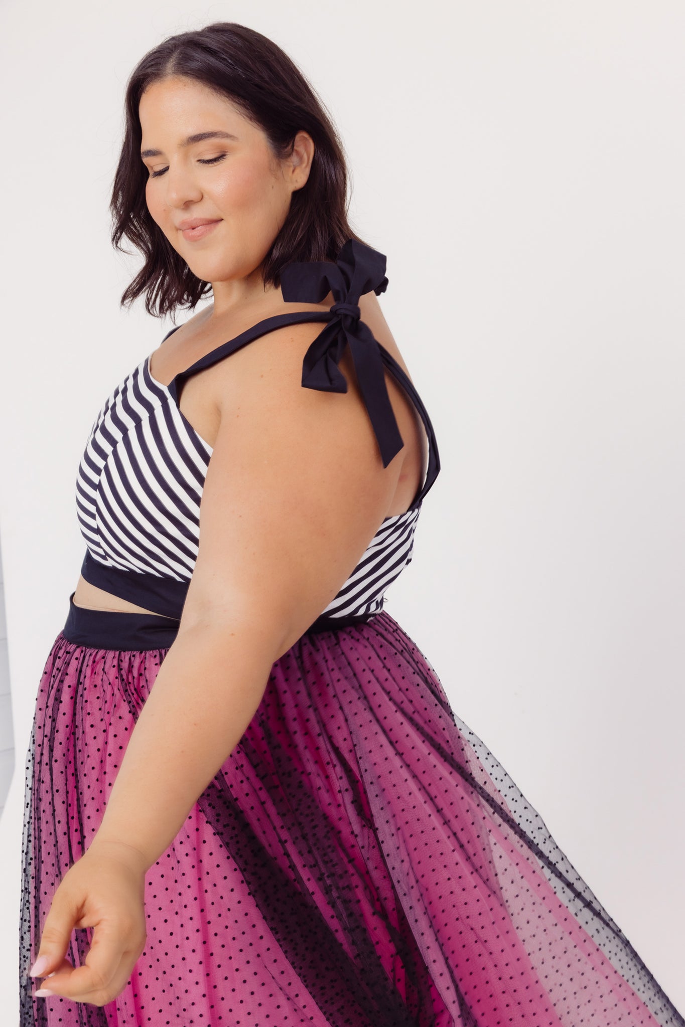 Side view of a plus size woman wearing a Cherry Bobbins black and white striped top with a bow and a pink polka dot tulle skirt
