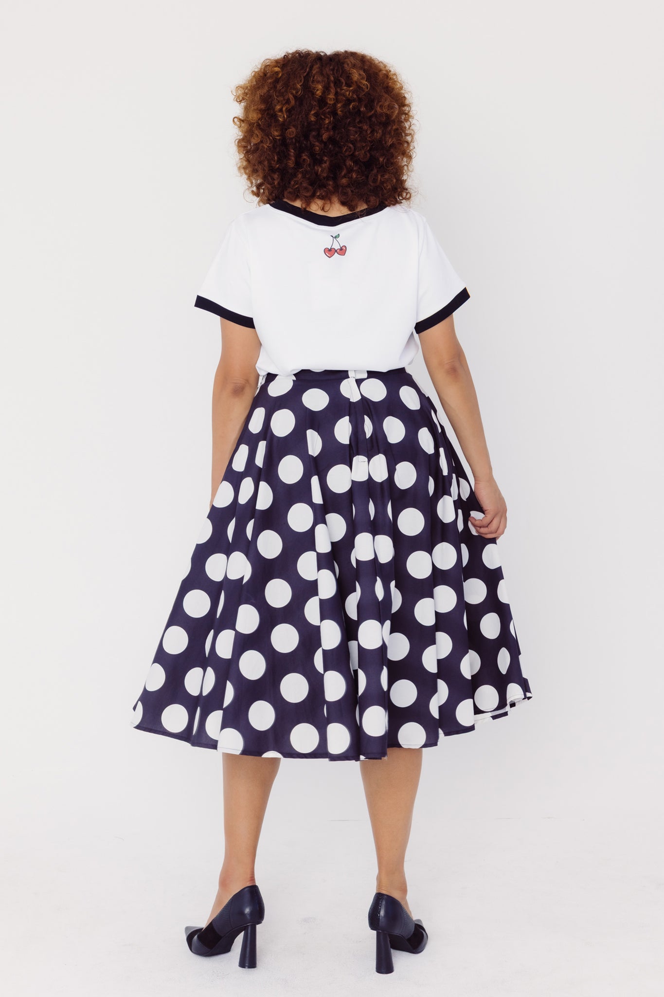Back view of a woman wearing a Cherry Bobbins white tee with black trim and a black polka dot cotton skirt on a white background