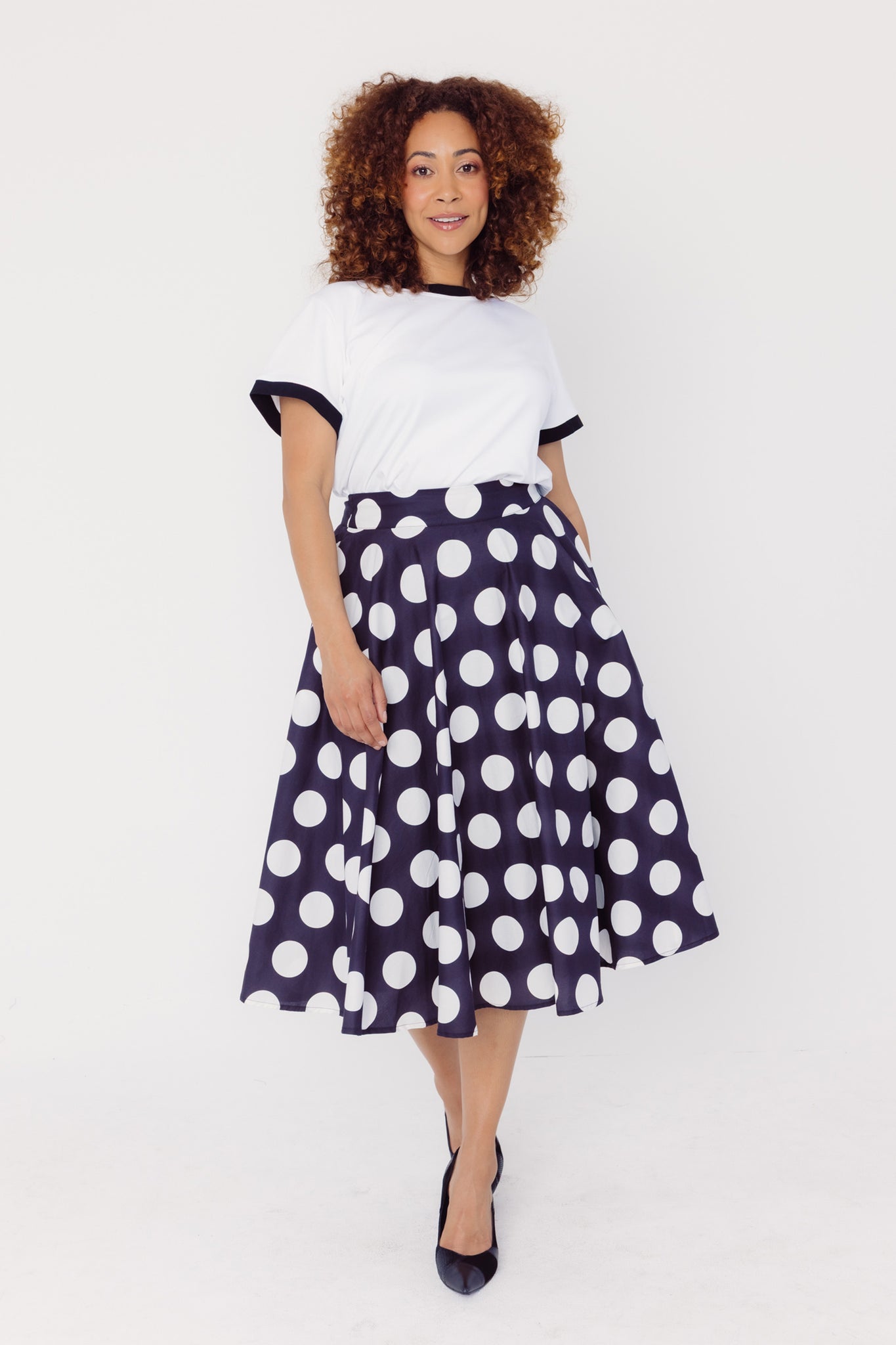 Front view of a woman wearing a Cherry Bobbins white tee and black polka dot cotton skirt on a white background