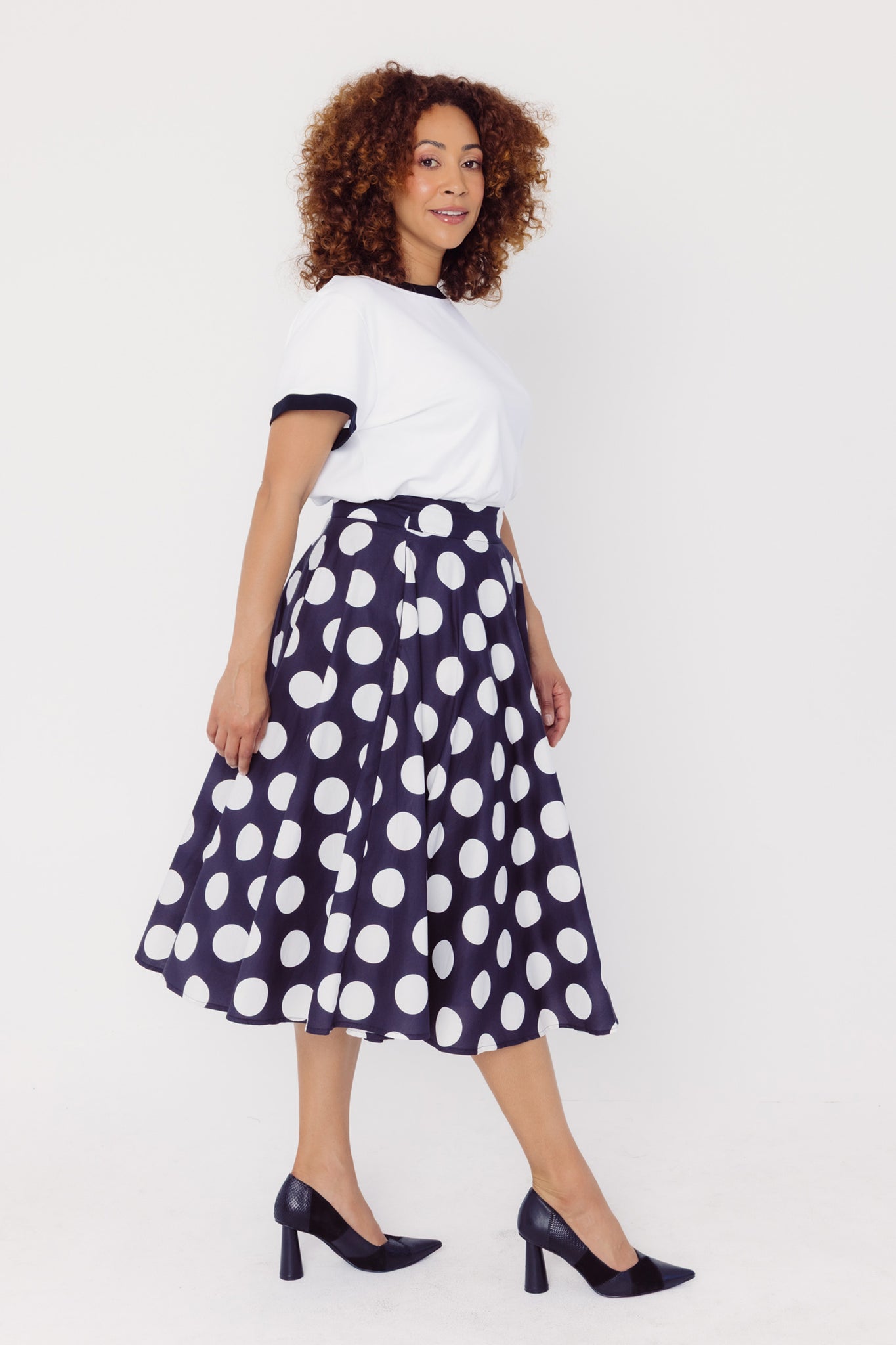 Side view of a woman wearing a Cherry Bobbins white t-shirt with black trim and a black cotton skirt with white polka dots on a white background