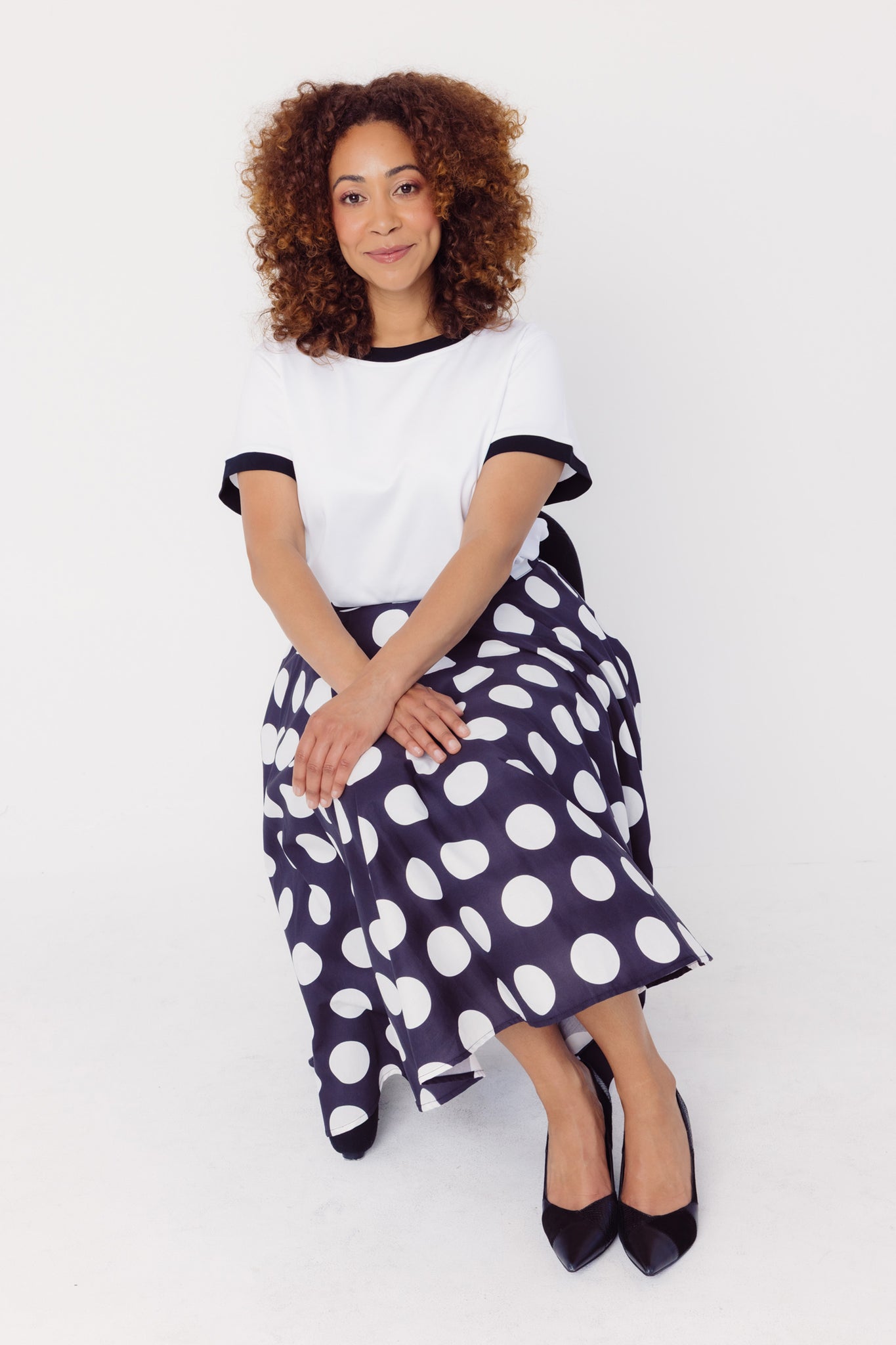 View of a woman wearing a Cherry Bobbins white t-shirt and polka dot skirt sitting on a white background