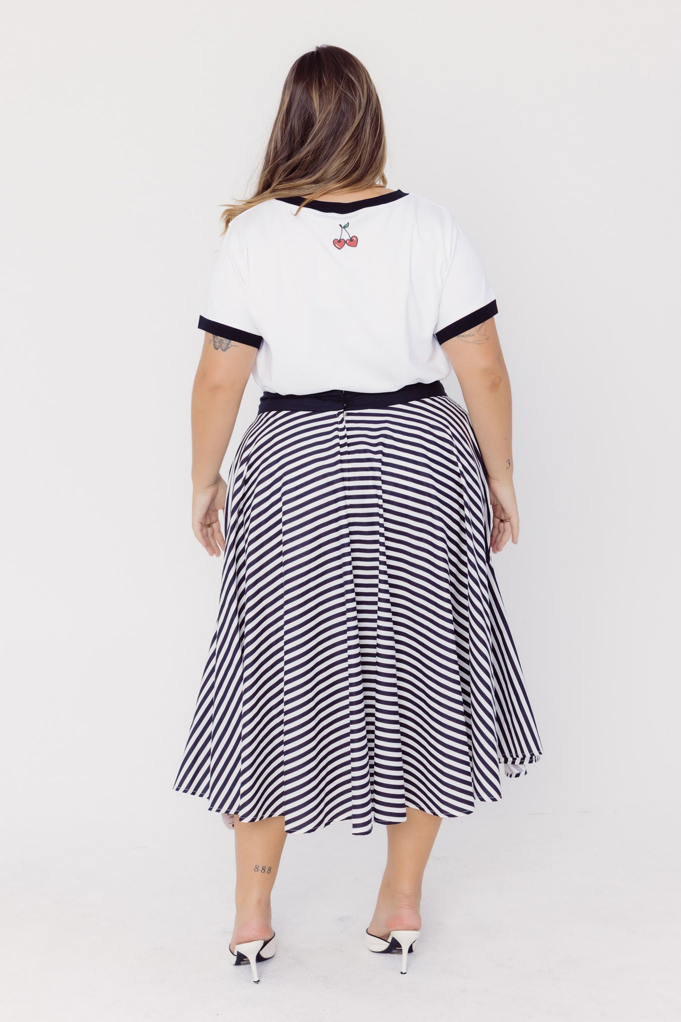 Back view of a woman wearing a Cherry Bobbins white t-shirt with black trim and a black and white striped cotton circle skirt on a white background