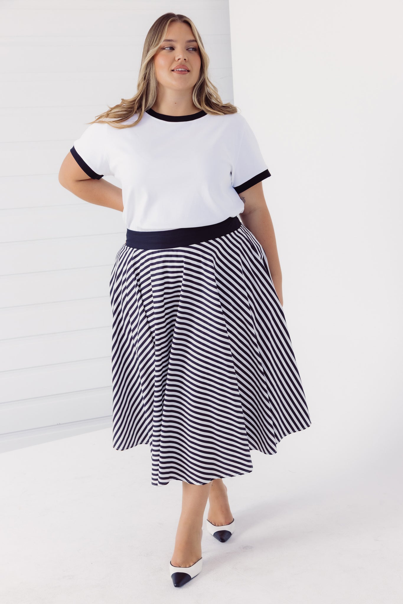 Full length view of a woman wearing a Cherry Bobbins white t-shirt with black trim and a black and white striped cotton circle skirt on a white background