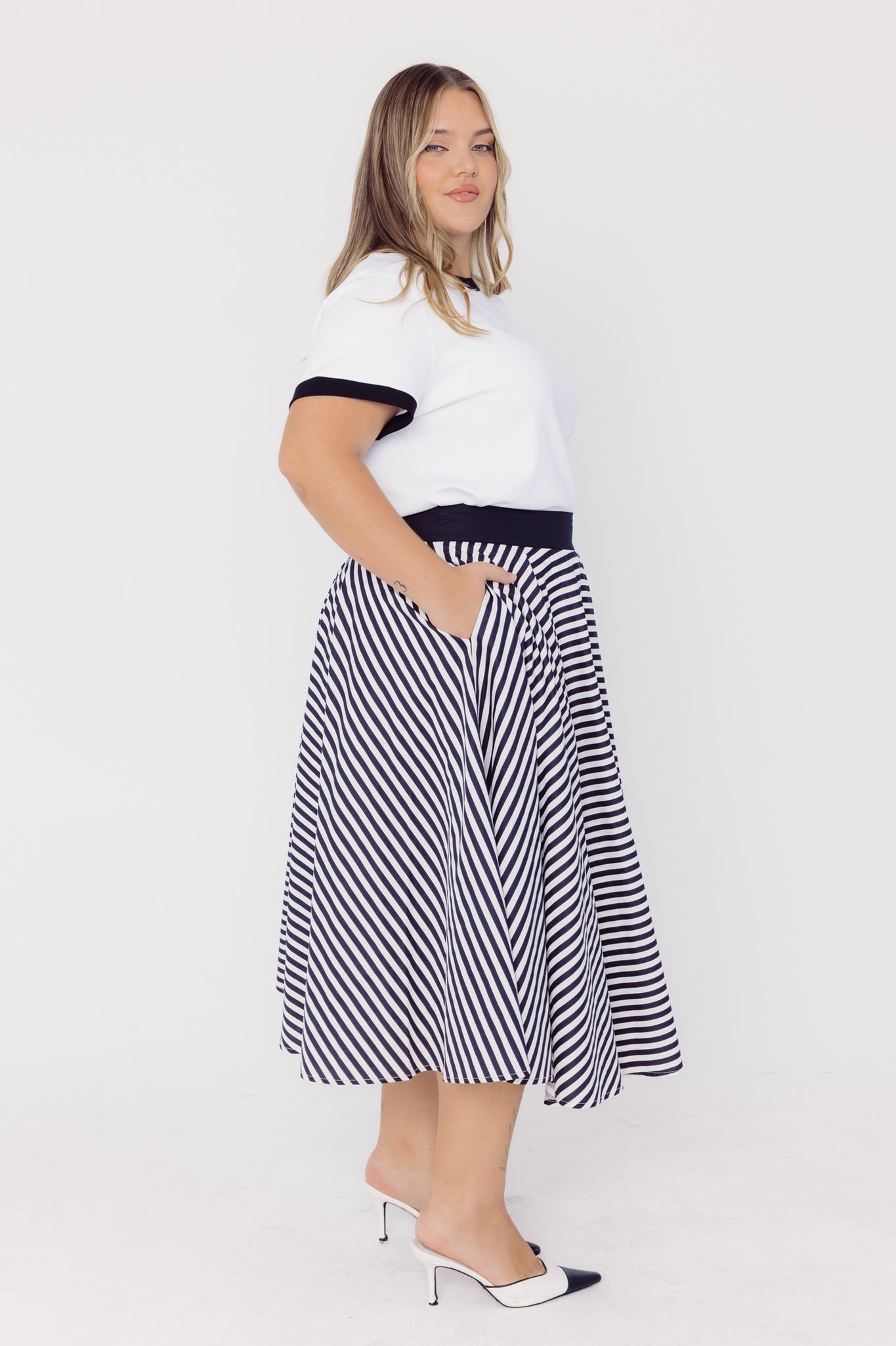 Side view of a woman wearing a Cherry Bobbins white tee with black trim and a black and white striped cotton circle skirt on a white background