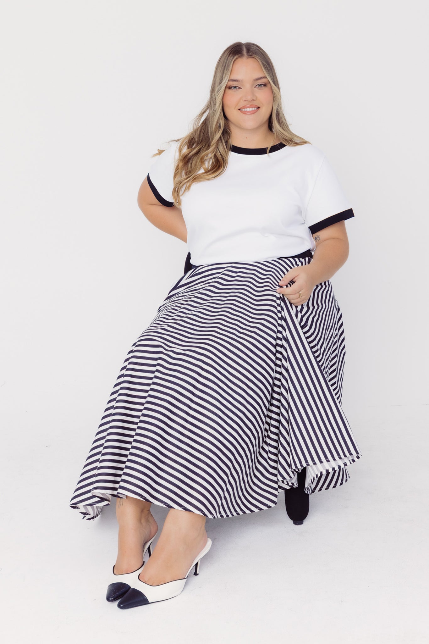 Woman wearing a Cherry Bobbins white T-Shirt with black trim and a black and white striped cotton skirt sitting on a chair