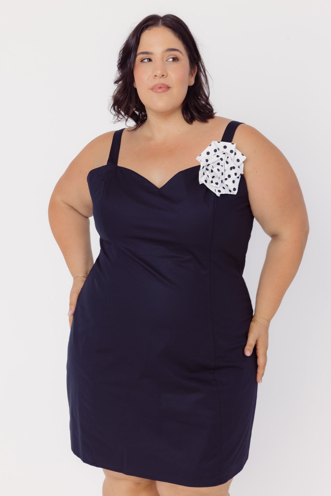 Woman wearing a black cotton slip dress with a white polka dot rose brooch detail on a white background