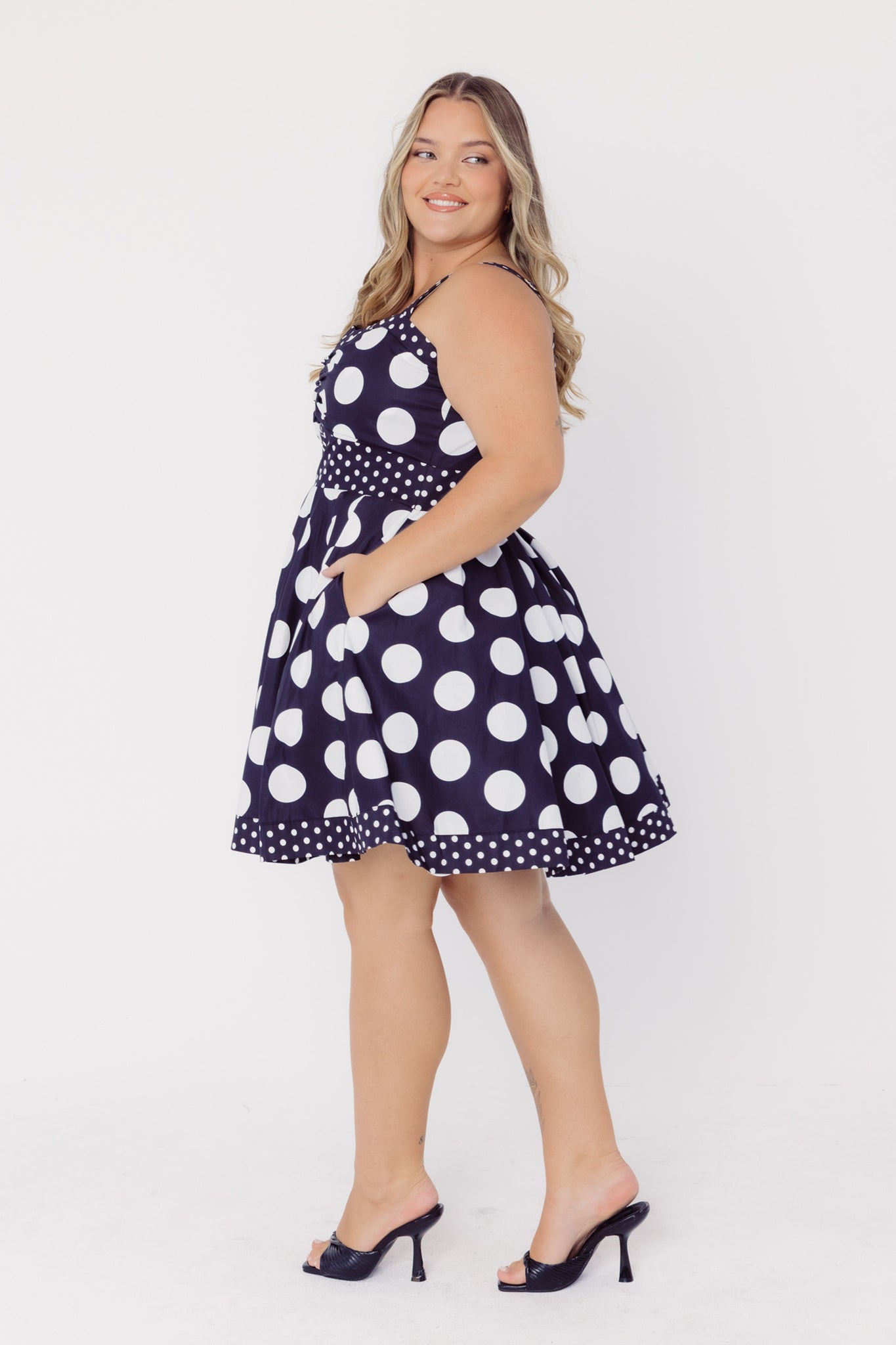 Side view of a woman wearing a Cherry Bobbins cotton mid-size black dress with white polka dots on a white background