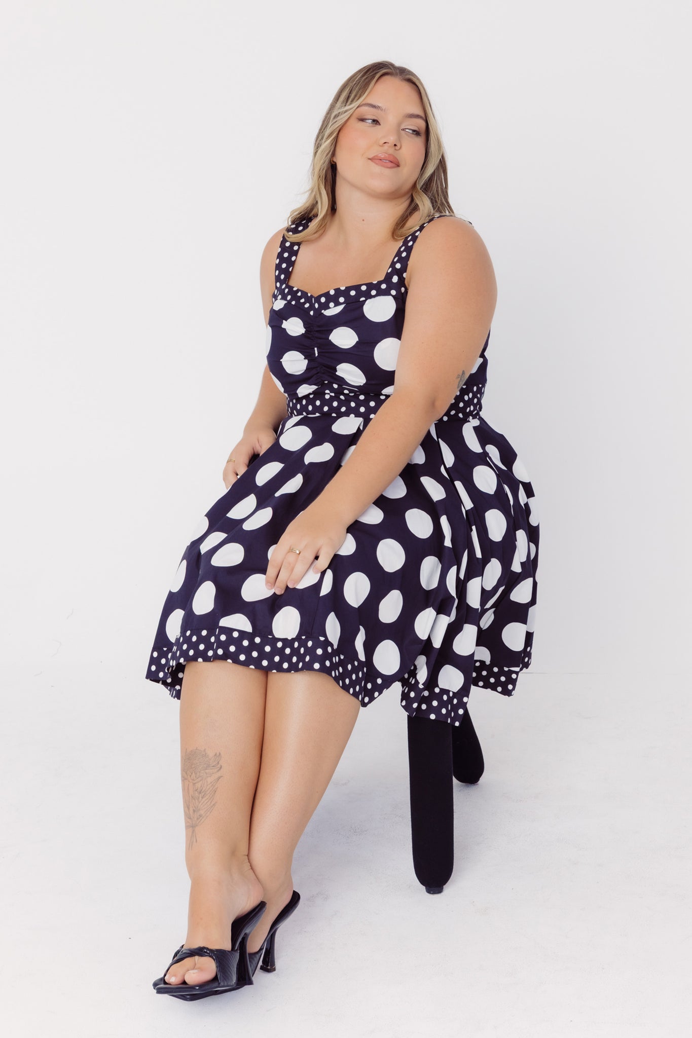 Woman sitting on a chair wearing a Cherry Bobbins black cotton mid-size dress with white polka dots on a white background