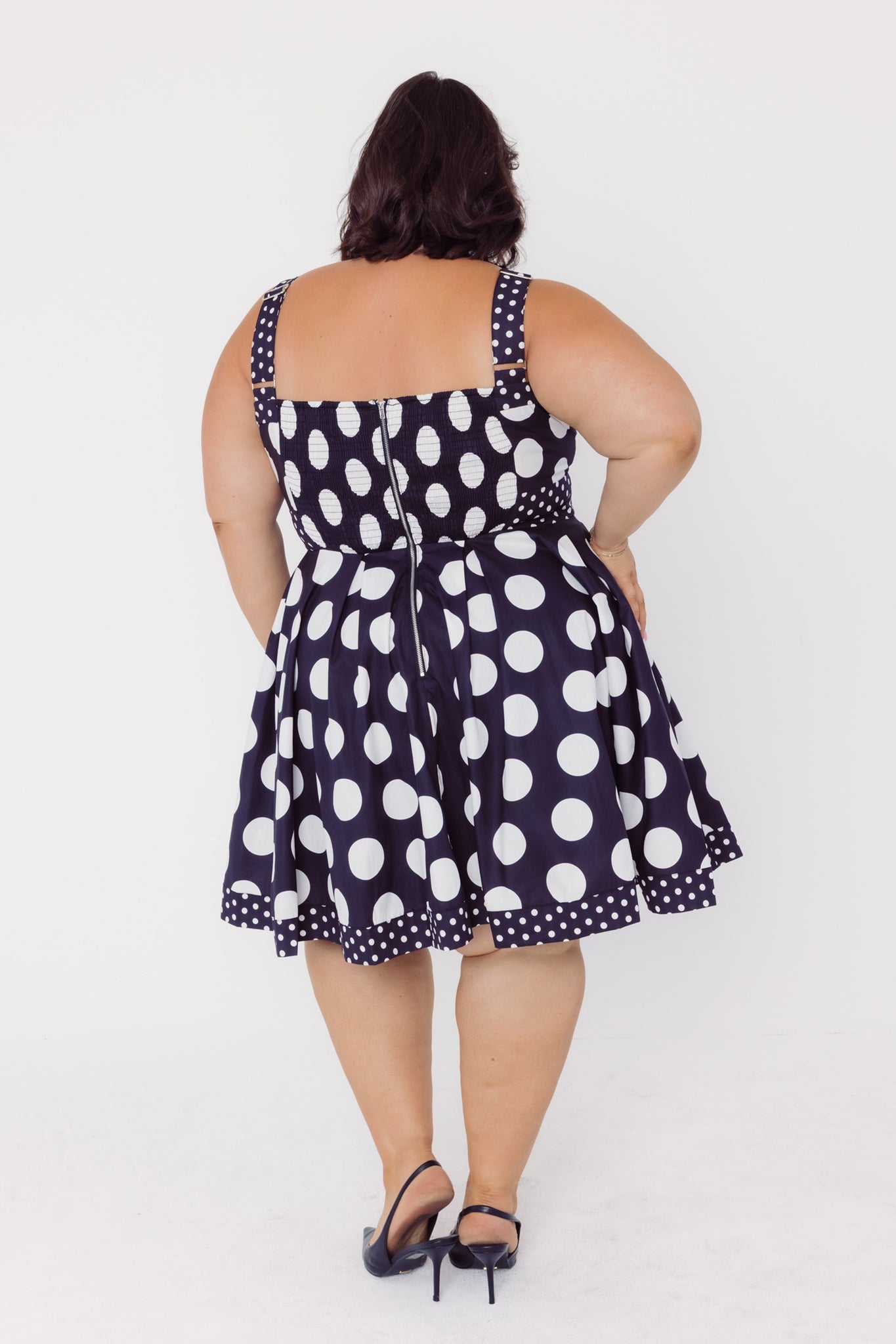 Back view of a woman wearing a Cherry bobbins black cotton polka dot dress on a white background
