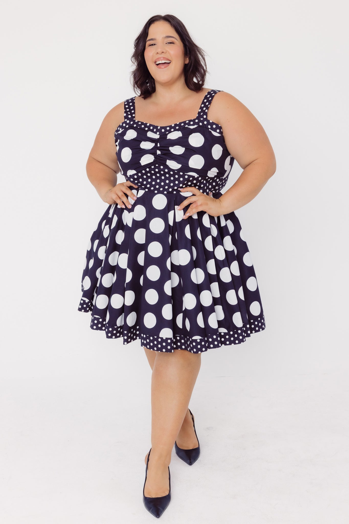 Full length front view of a woman wearing a Cherry Bobbins black cotton plus size dress with white polka dots on a white background