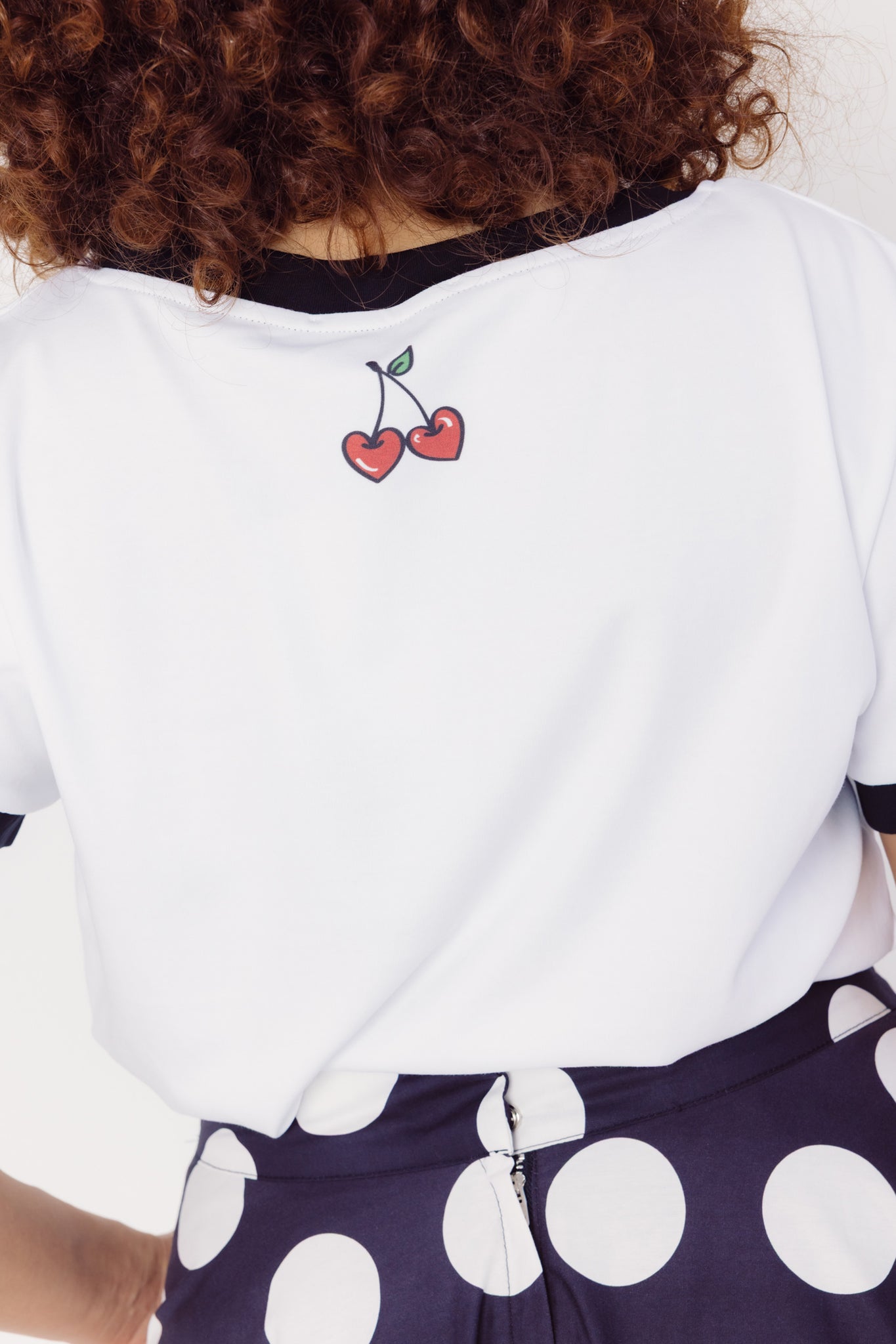 Woman wearing a white cotton blend t-shirt with cherry design on the back and polka dot cotton skirt