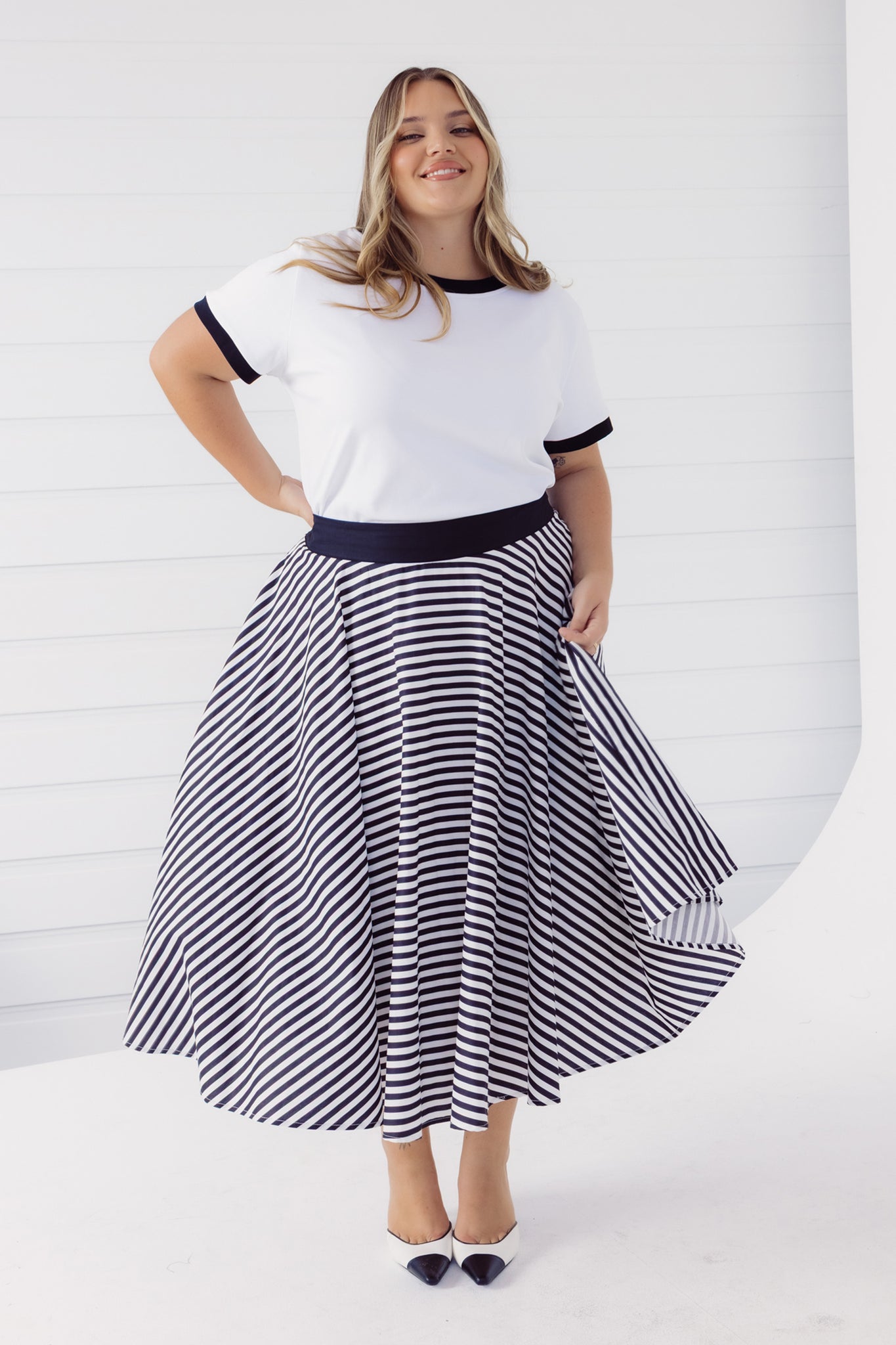 Full length view of a woman wearing a Cherry Bobbins white cotton blend t-shirt with black trim and a black and white striped skirt on a white background
