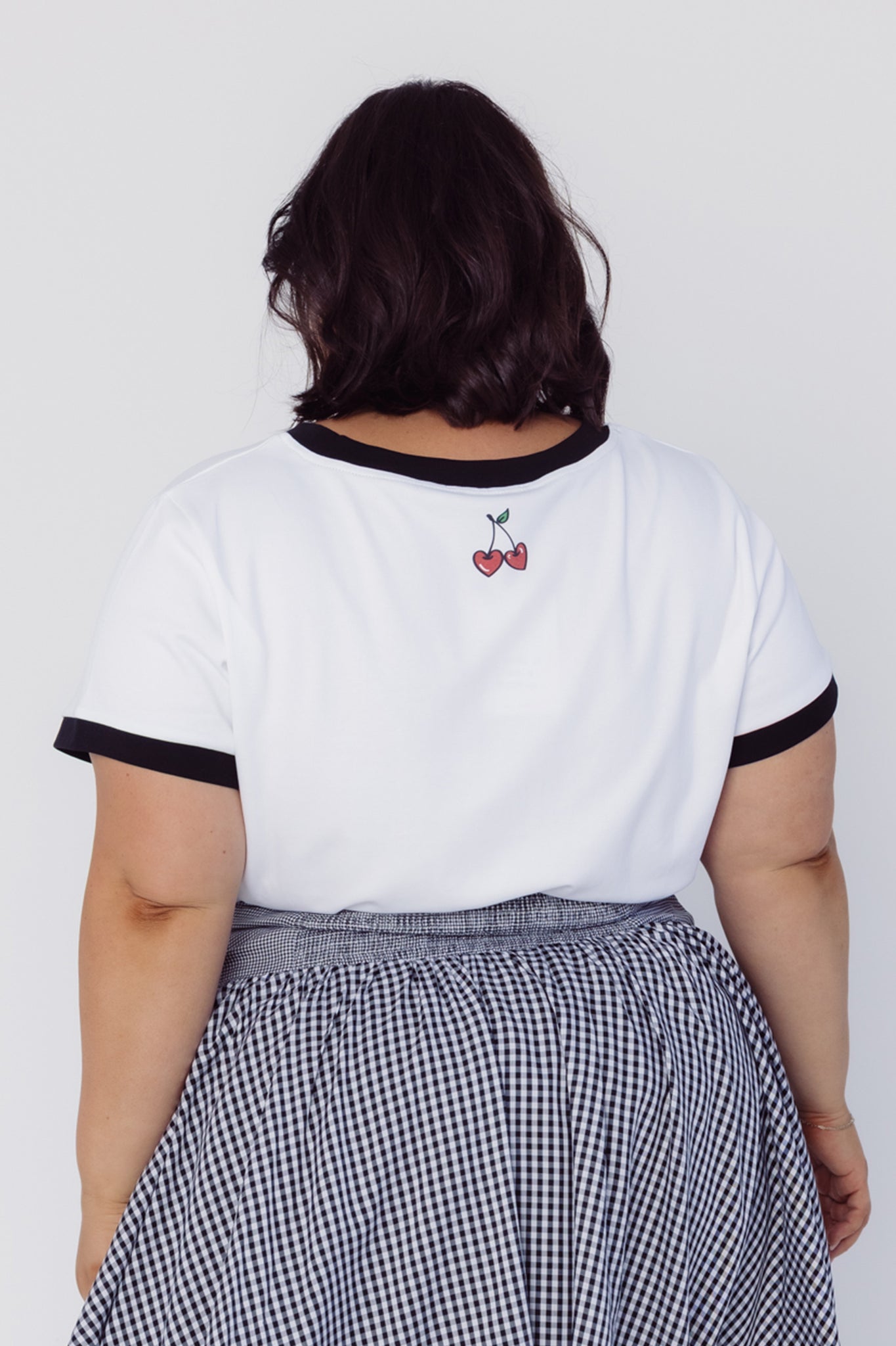 Back view of a woman wearing a Cherry Bobbins white t-shirt with black trim and a checkered skirt on a plain background