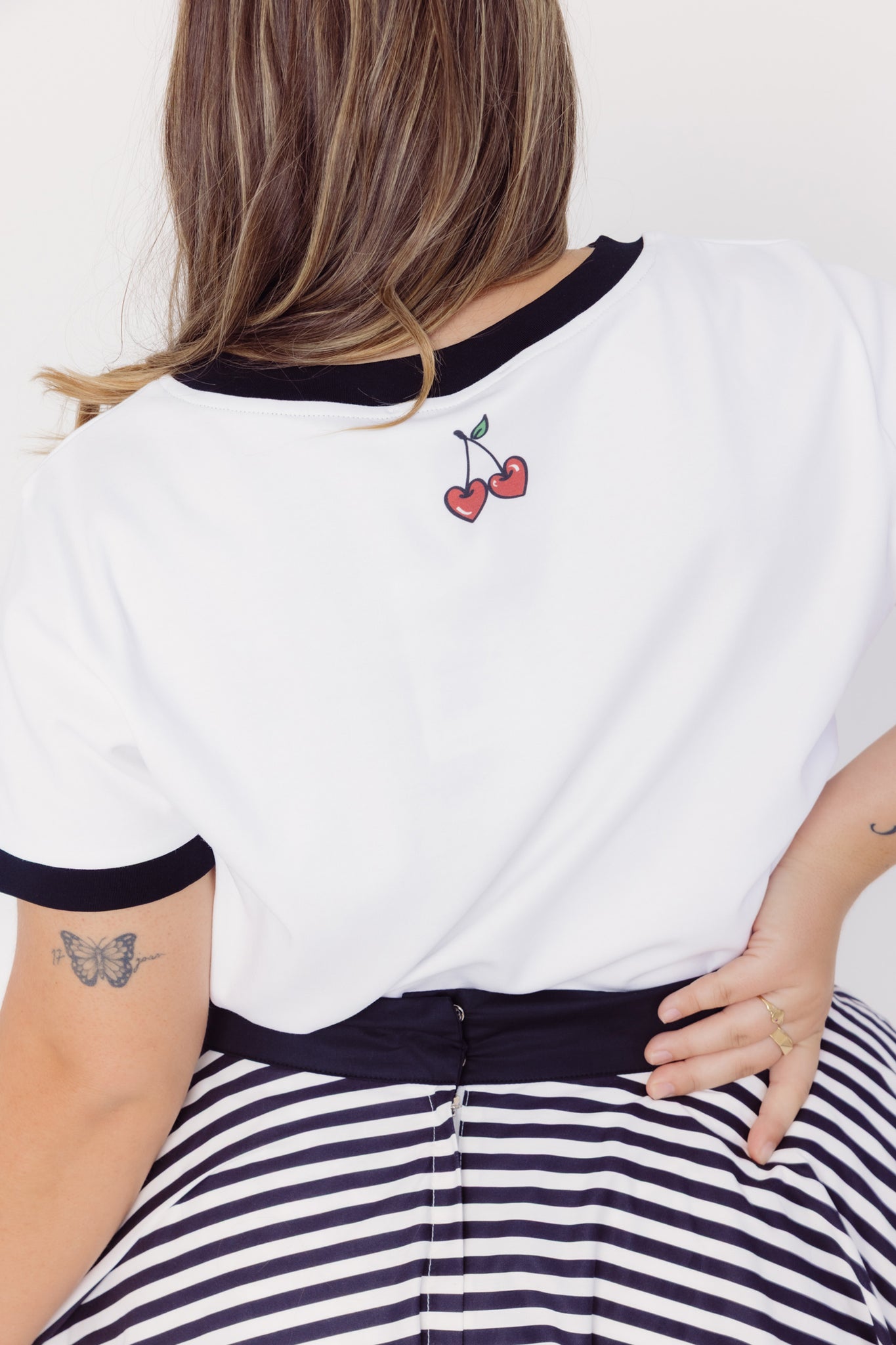 Back close-up view of a woman wearing a Cherry Bobbins white cotton blend t-shirt with black trim and a cherry design, paired with a cotton striped skirt.