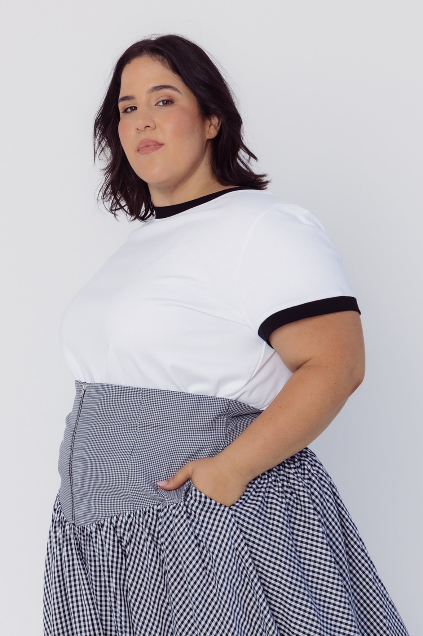 Side view of a woman wearing a Cherry Bobbins white t-shirt with black trim and a checkered skirt on a plain background