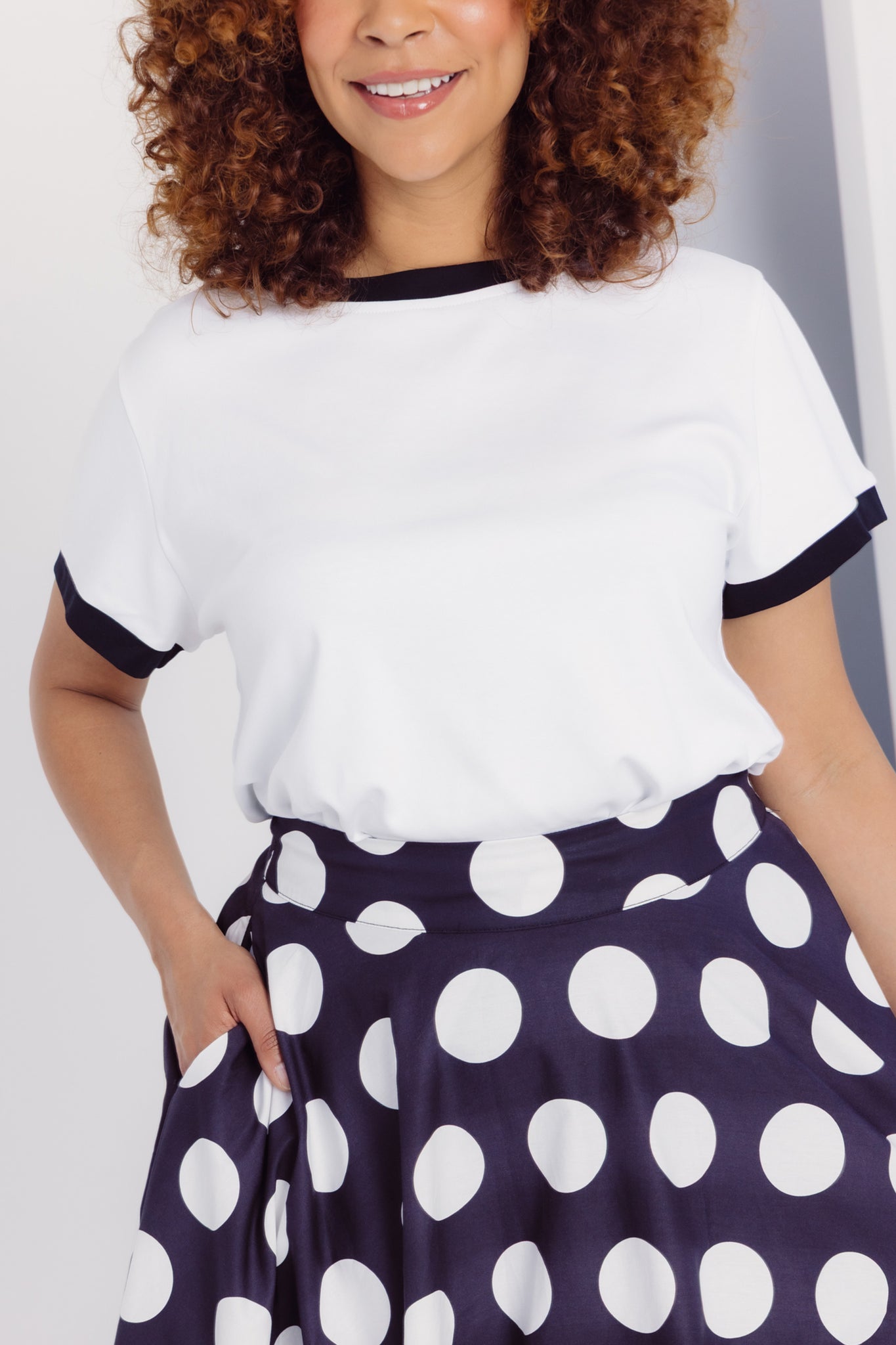 Close up view of a woman wearing a Cherry Bobbins white cotton blend t-shirt with black trim and a black skirt with white polka dots.