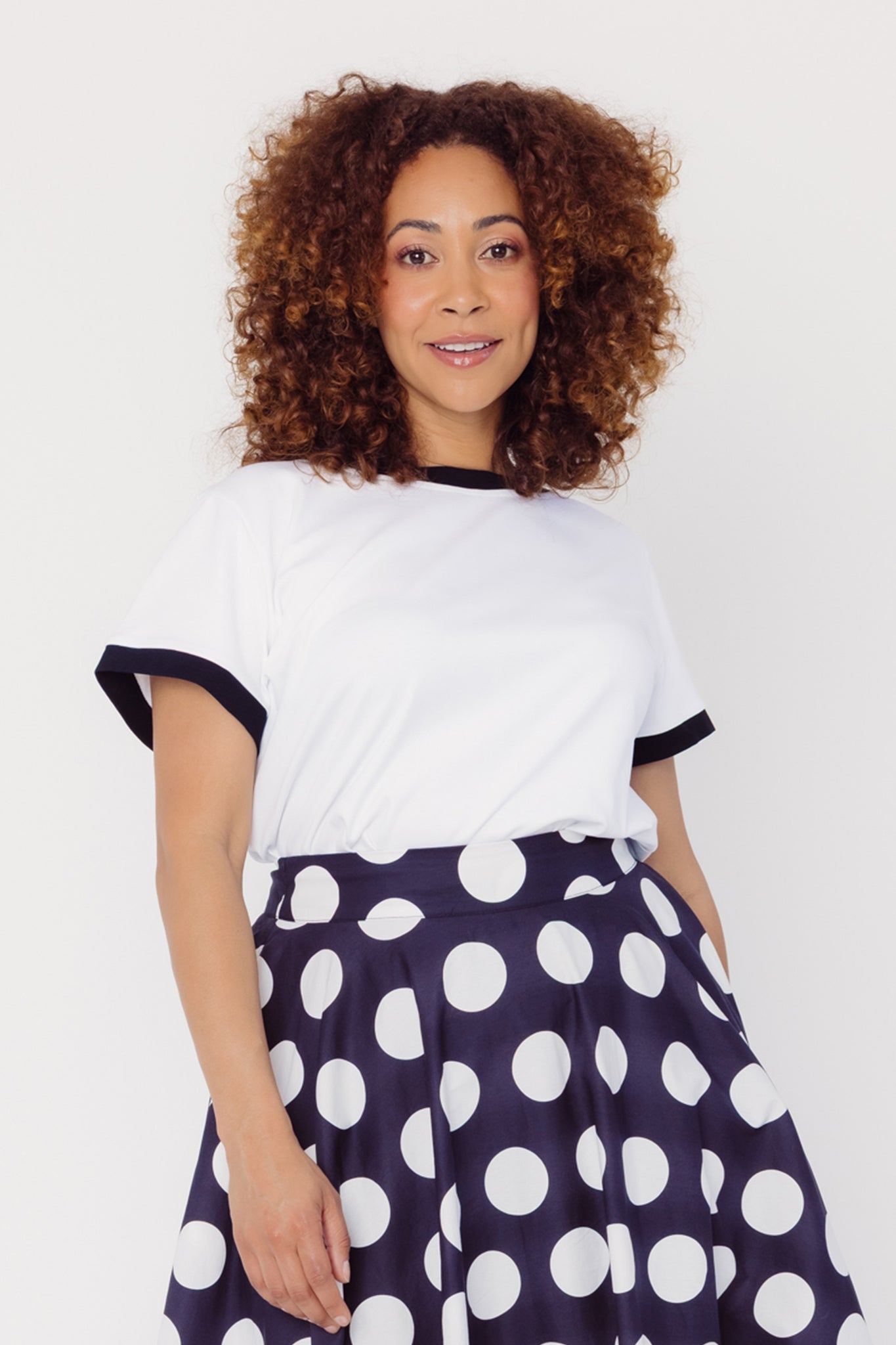 Front view of a woman wearing a Cherry Bobbins white cotton blend top with black trim and a black skirt with white polka dots on a plain background
