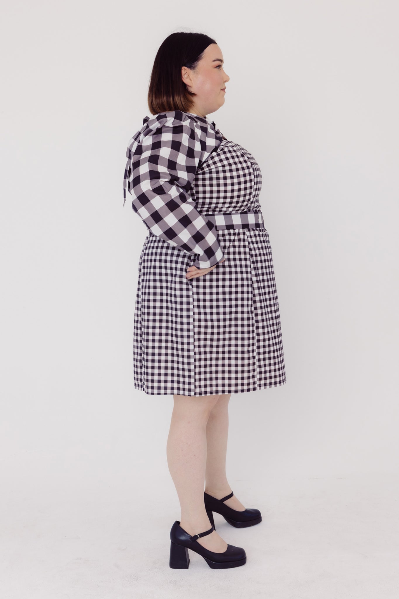 Side view of a woman wearing a black and white Cherry Bobbins checkered plus size dress on a white background