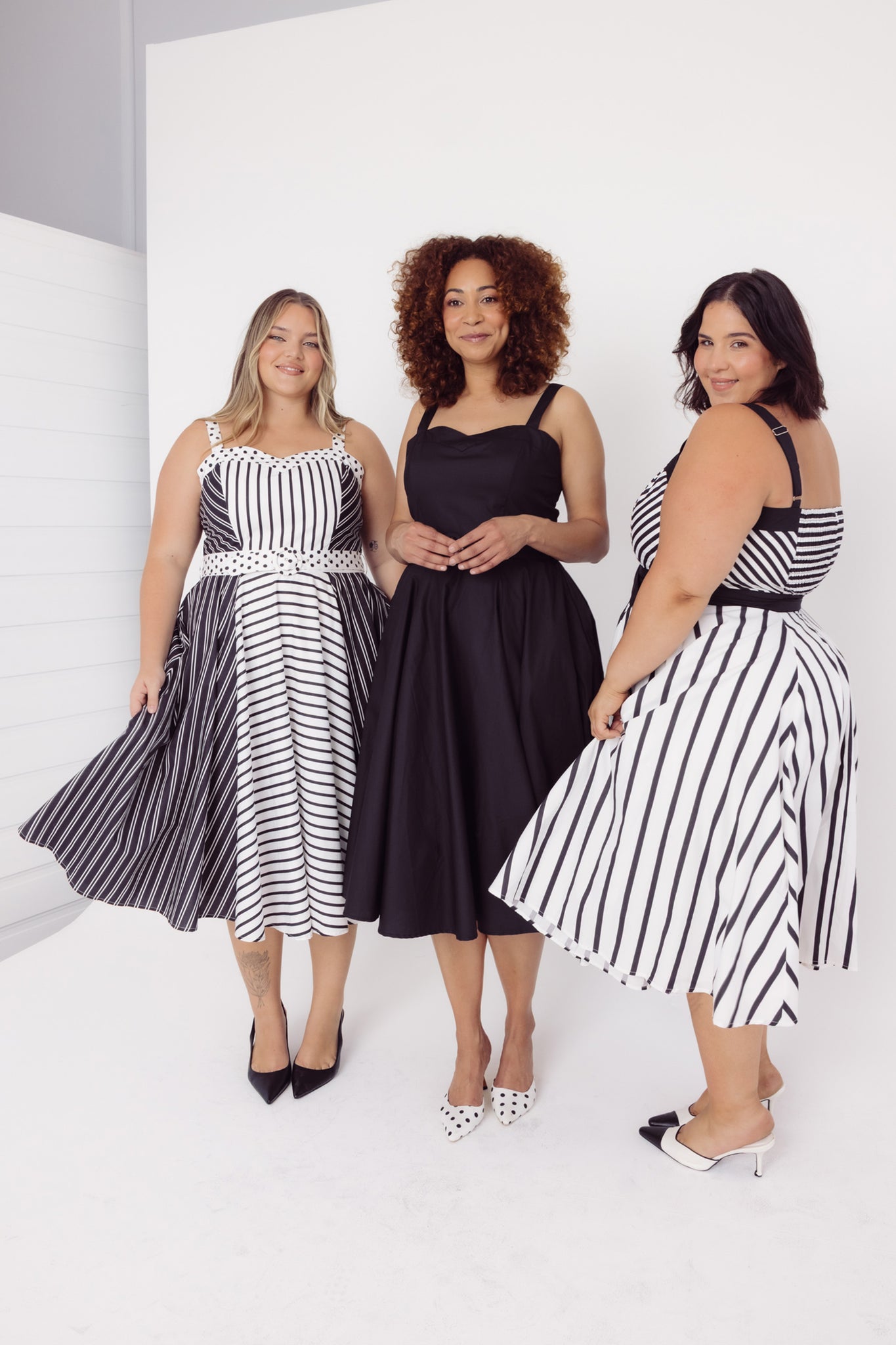Three women wearing Cherry Bobbins black and white striped and plain midi cotton dresses on a white background