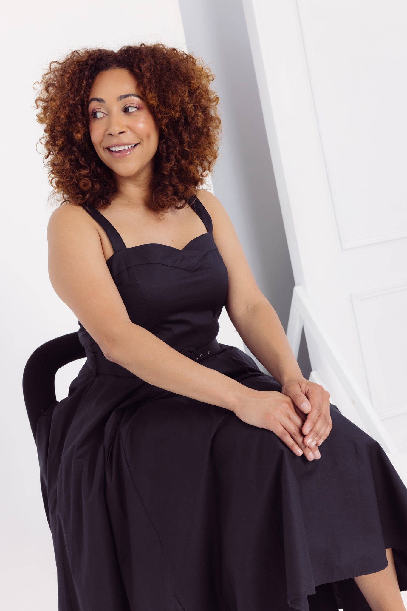 Close-up view of a woman in a Cherry Bobbins plain black cotton dress sitting on a chair against a white background
