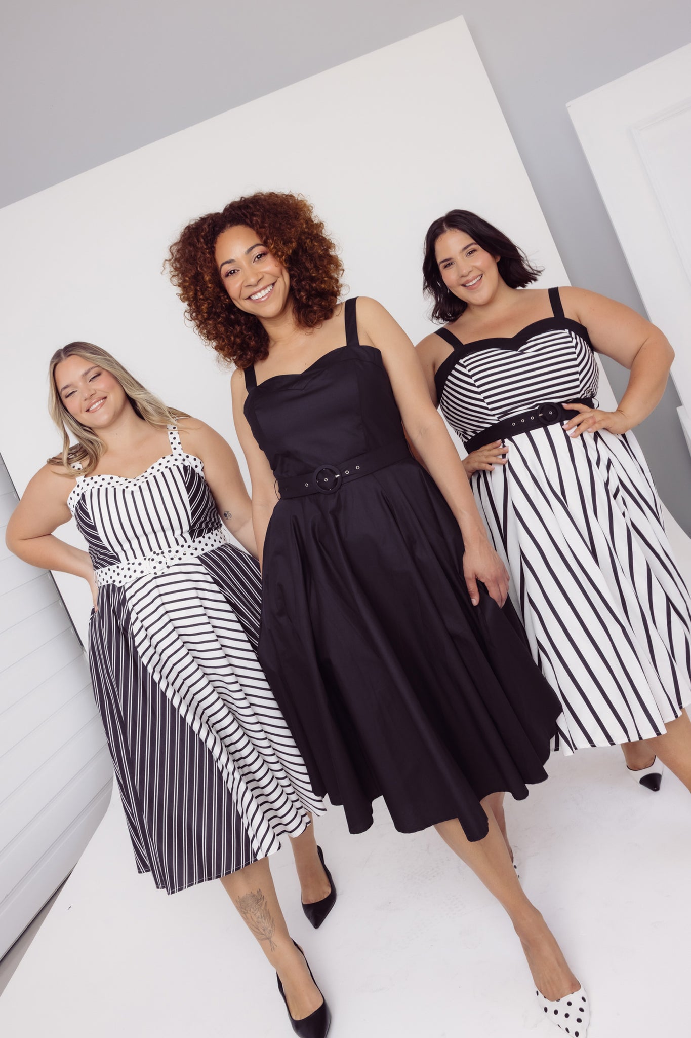 Three women wearing Cherry Bobbins black and white striped cotton midi dresses strutting in a studio and posing against a light gray background.