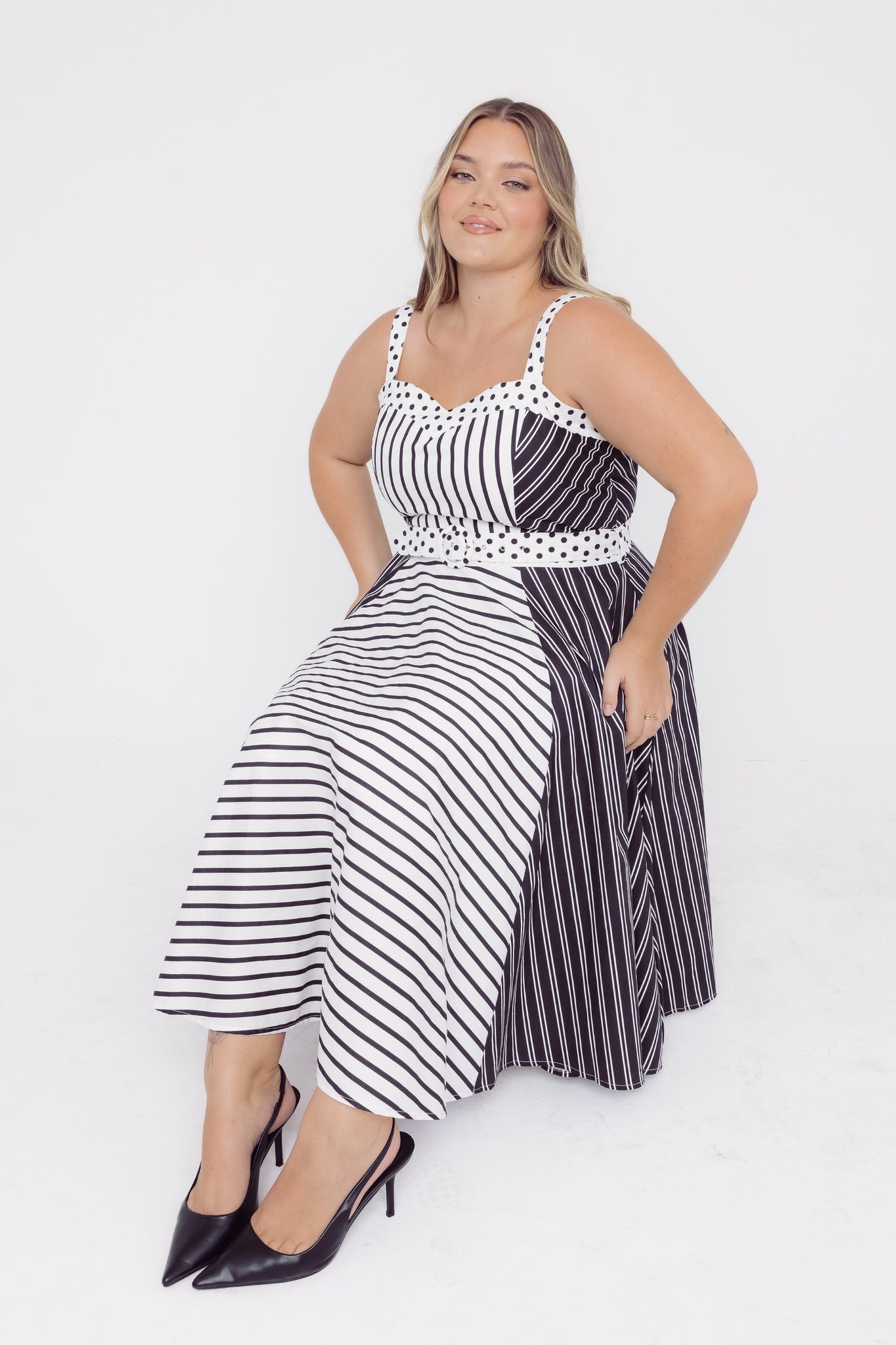 Mid-sized woman sitting on a chair wearing a Cherry bobbins black and white striped dress with a polka dot trim and self covered belt
