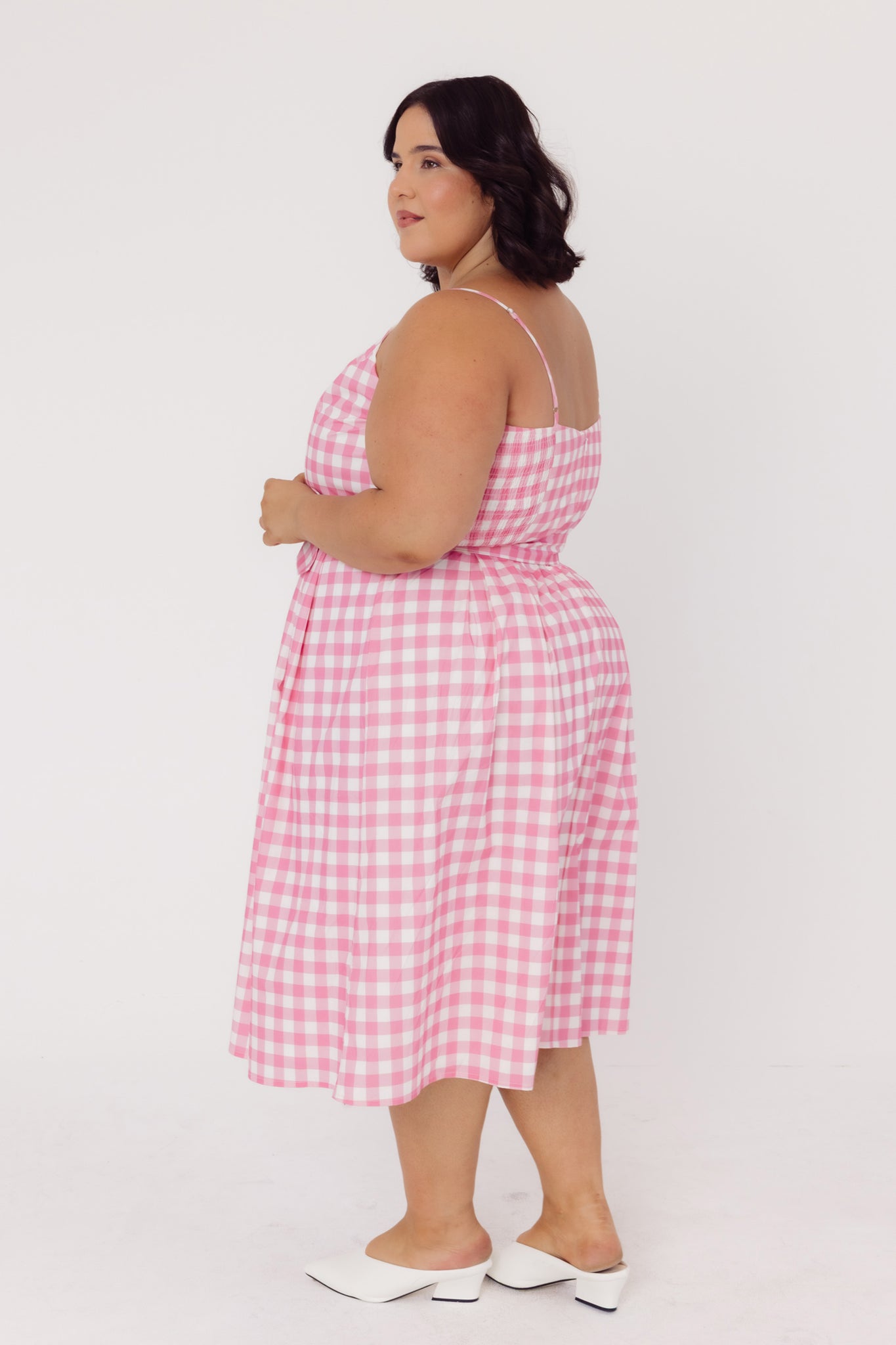 Back side view of a woman wearing a Cherry Bobbins sustainable cotton pink gingham dress on a white background