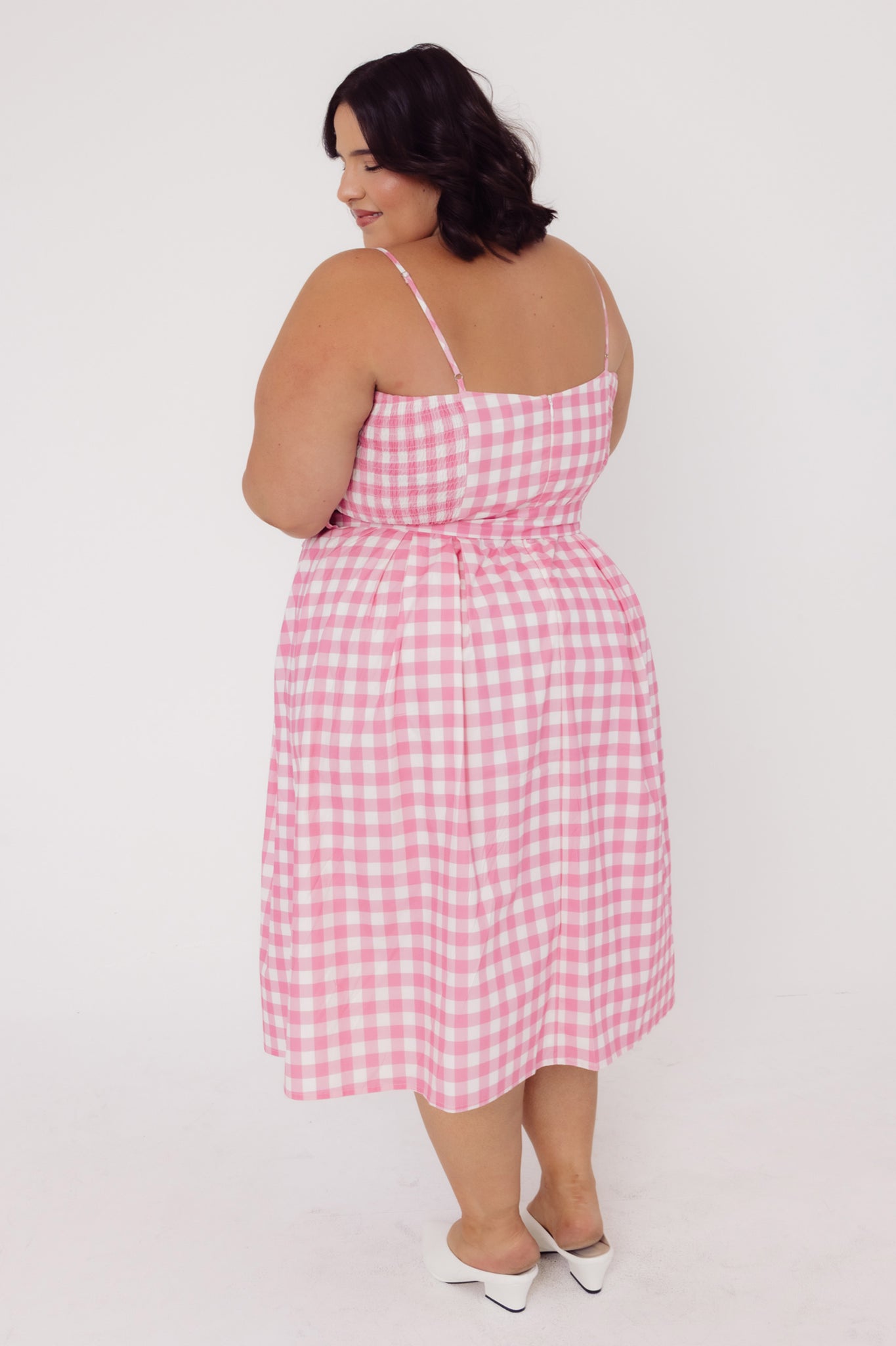 Back view of a woman wearing a Cherry Bobbins pink cotton gingham dress on a white background