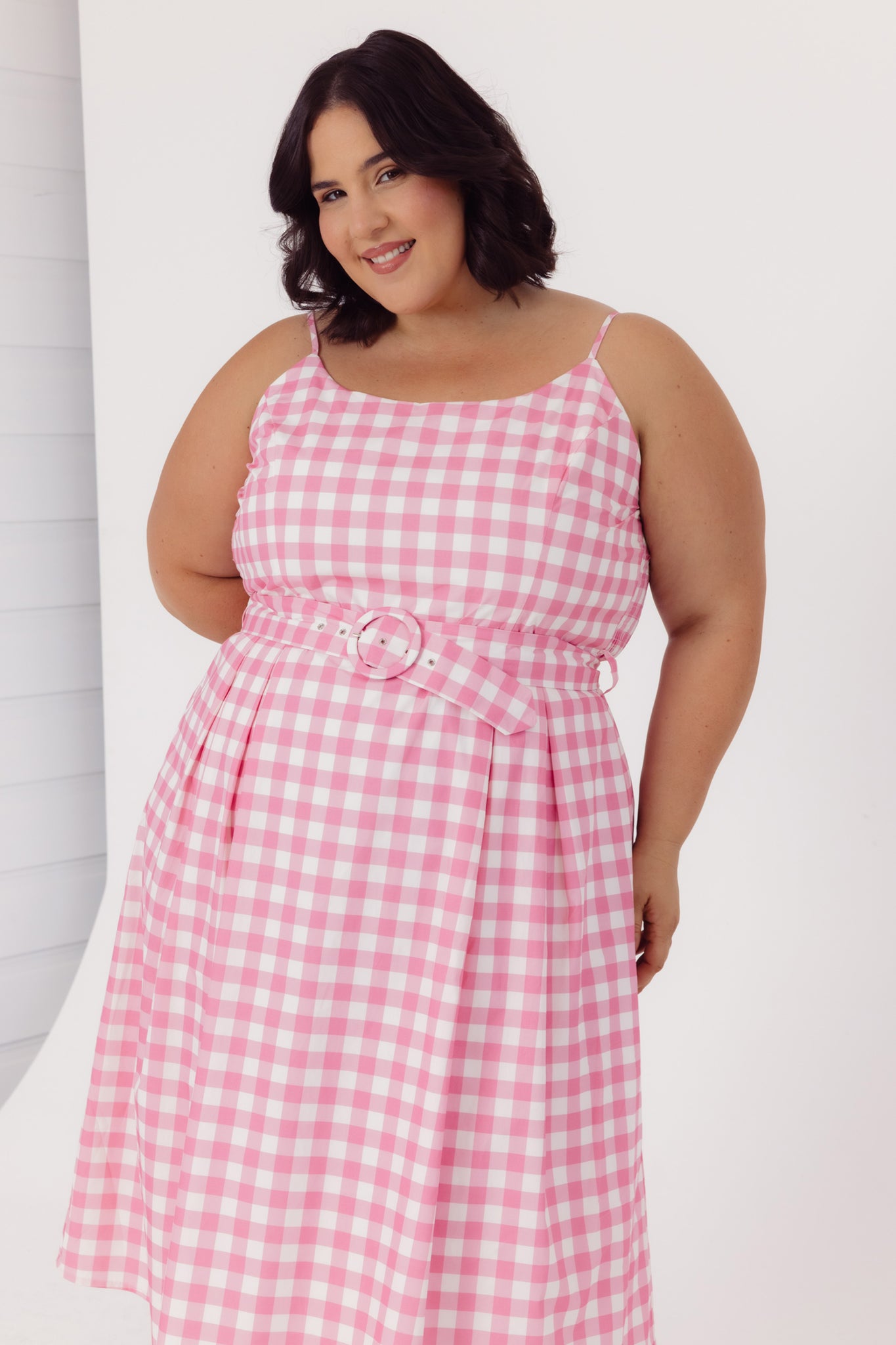 Plus size woman wearing a Cherry Bobbins sustainable cotton pink gingham dress with a belt on a white background