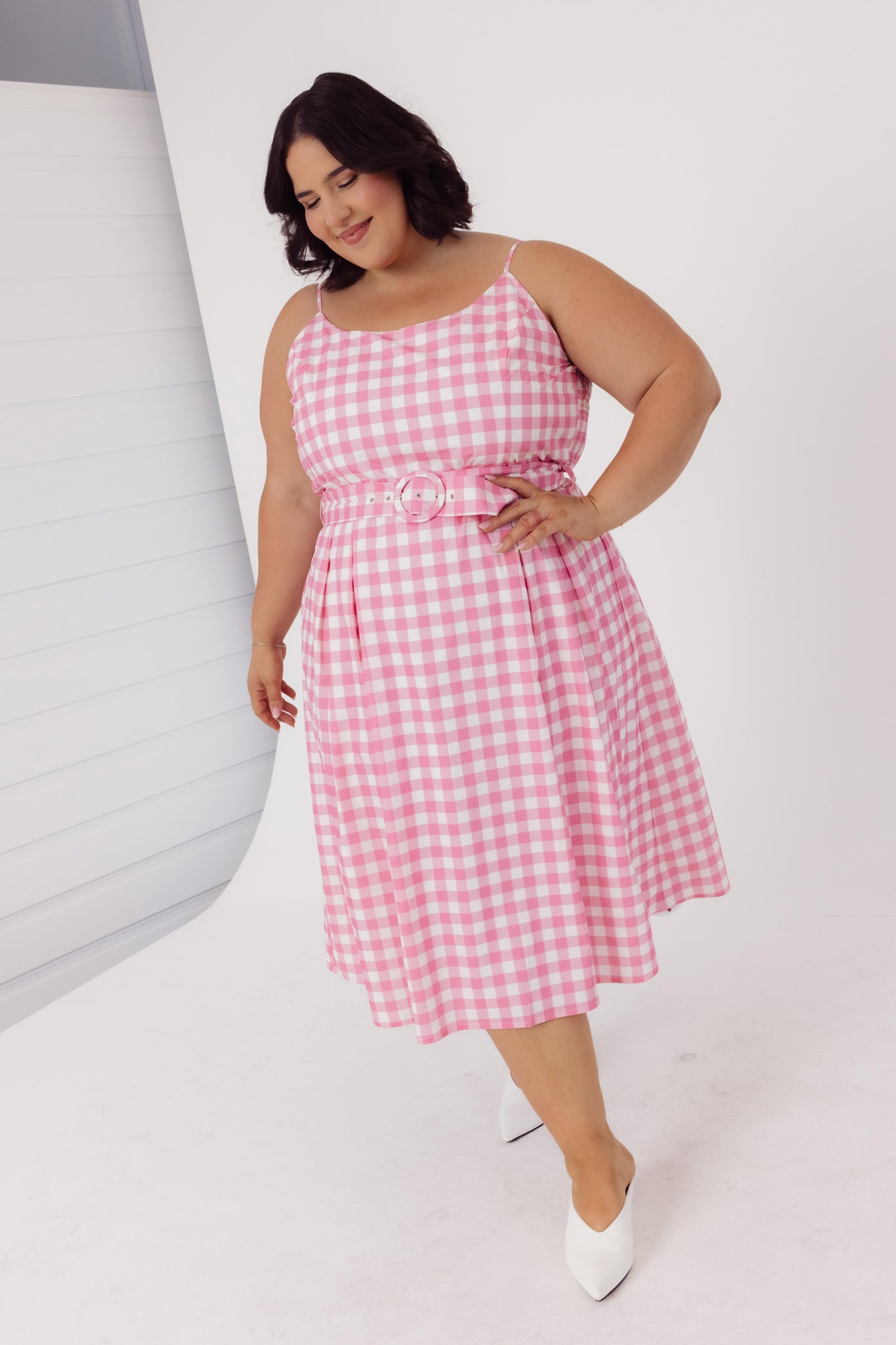 Full length view of a plus size woman wearing a Cherry Bobbins pink sustainable cotton gingham dress in a studio photoshoot