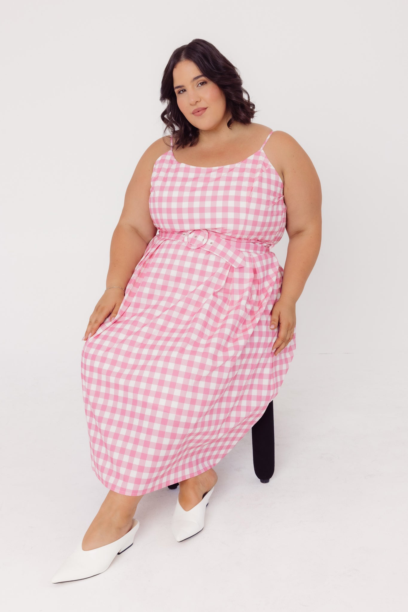 Woman sitting on a black chair wearing a Cherry Bobbins sustainable cotton pink gingham dress on a white background