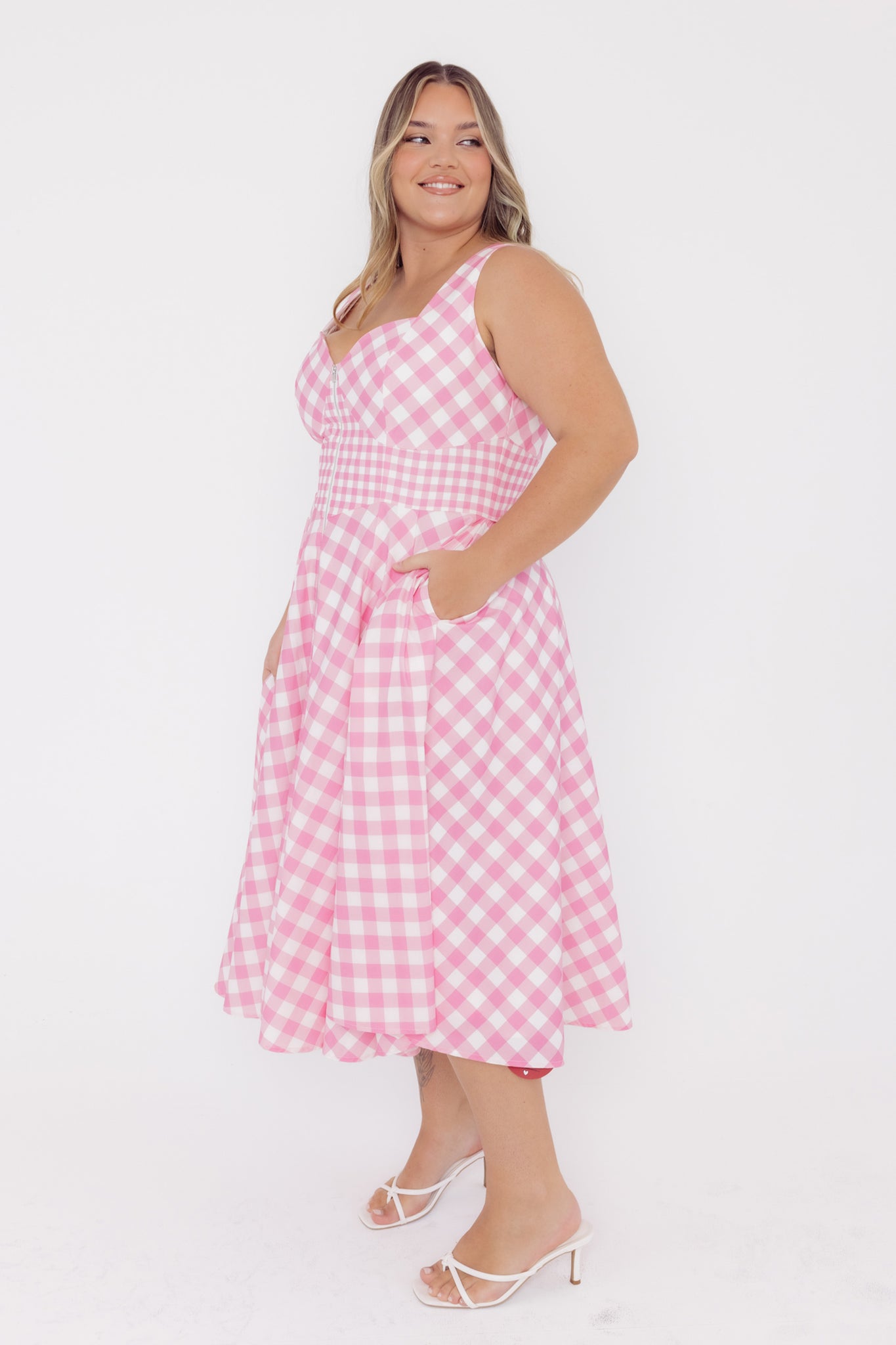 Side view of a woman wearing a Cherry Bobbins pink and white gingham midi cotton dress on a white background