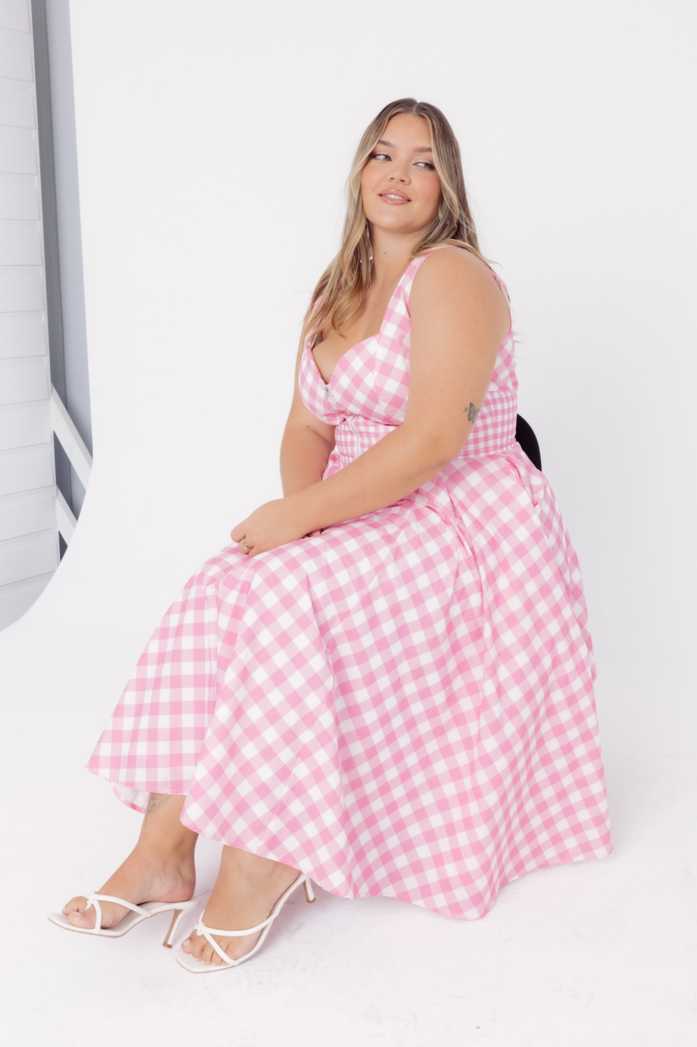 Side view of a woman wearing a Cherry Bobbins pink gingham midi cotton dress sitting on a black chair