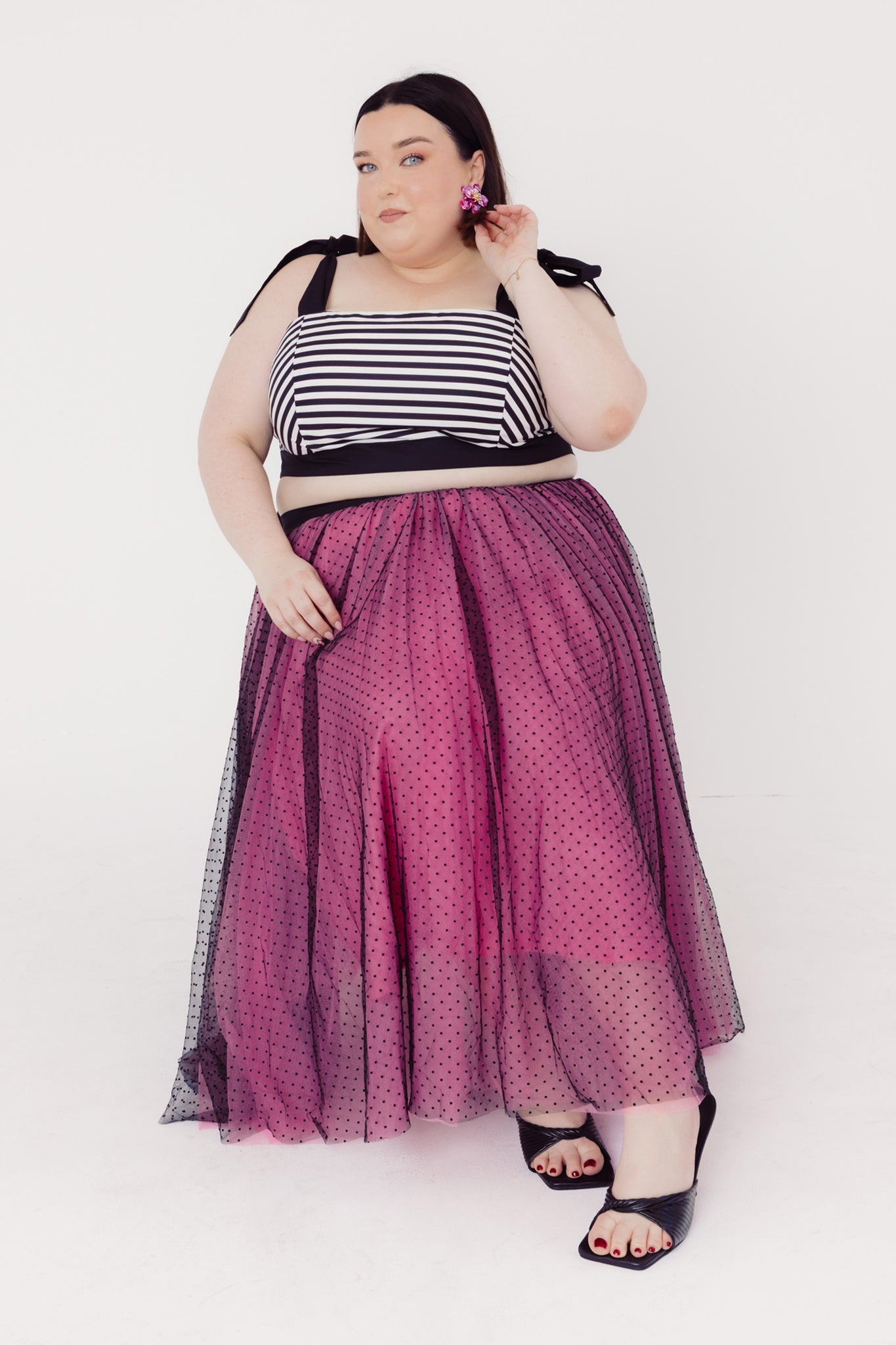 Woman sitting on a chair wearing a Cherry Bobbins black and white striped cotton top with a pink tulle polka dot skirt on a white background