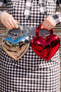 Person holding two metallic heart-shaped bags, one silver and one red, against a checkered dress