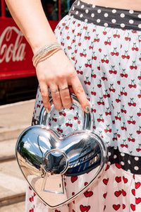 Person holding a silver heart-shaped handbag with a Cherry bobbins multi patterned skirt.