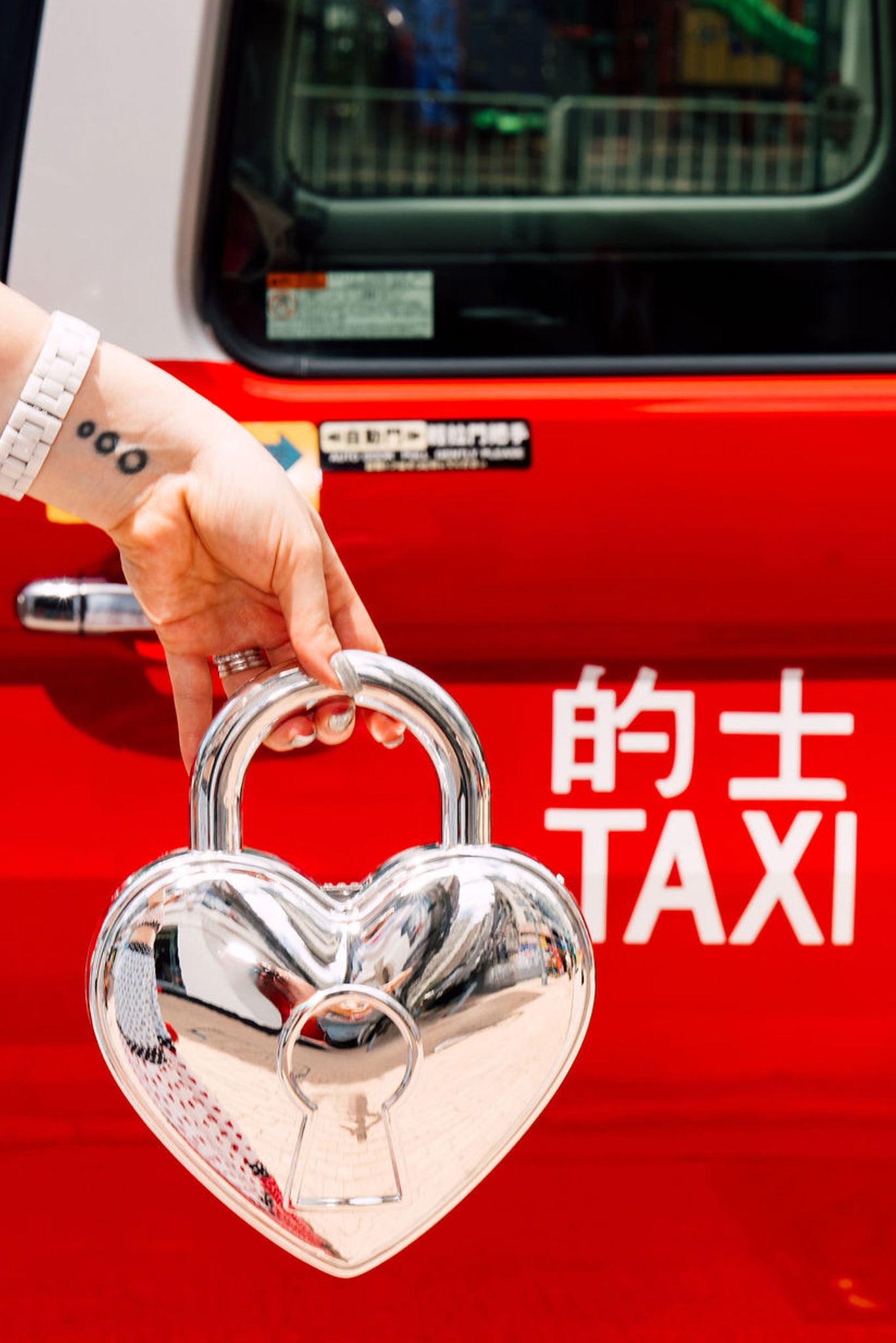 Heart-shaped locket bag held by a hand in front of a red taxi in Hong Kong