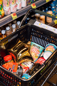 Shopping cart filled with various products including snacks, drinks and Cherry Bobbins gold star bag, in a grocery store setting.